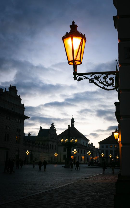 A street lamp glows warm orange in the twilight showing the Gothic castle square of Prague, Czechia